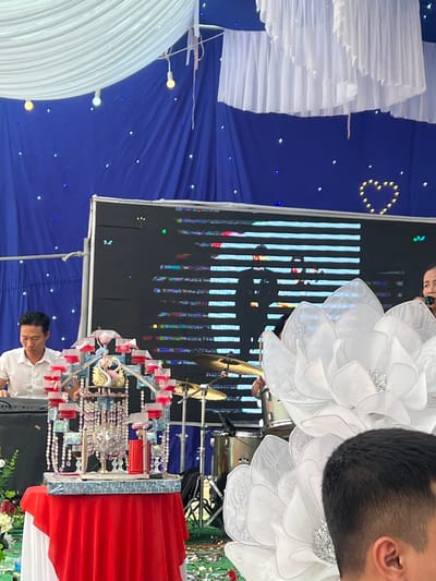 A horizontal indoor shot shows decorations and people at a ceremony in Tay Son Commune, Vietnam. A blue backdrop with twinkling lights forms the background. Decorations include large white floral arrangements, white draped elements, and an elaborate beaded and bejeweled structure with red accents and golden swans. A digital screen displaying vertical lines and a silhouette of a person stands prominently. Two men are visible, one in a white shirt and one in the upper right corner with short black hair. The man in the white shirt is positioned near the beaded structure, while the other is partially obscured. The scene suggests a formal event with a blend of traditional and modern elements.