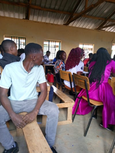 This image captures an indoor scene, likely a classroom or community hall, in Mwingi, Kenya. The room is populated by a group of people, primarily young adults, seated on simple furniture.
In the foreground, a young man with short dark hair, wearing a light blue polo shirt and grey jeans, sits on a rustic wooden bench. He is turned slightly to his right, looking away from the camera, with his hands clasped in front of him. His expression appears calm and attentive.
Behind him and to his right, several other individuals are seated. A woman with distinct long, voluminous red braids is seen from behind, wearing a white top. Next to her, another woman is partially visible, wearing a patterned top. Further to the right, a woman in a vibrant magenta or fuchsia dress, also with long black braided hair cascading down her back, sits facing away from the viewer on a chair with a wooden back and metal frame. Several other people are discernible in the background, mostly seen from behind or in profile.
The setting features light-colored, possibly unpainted, walls with multiple windows fitted with green-painted metal security grilles. The ceiling is made of corrugated metal sheets supported by exposed wooden beams. The furniture consists of basic wooden benches and individual chairs. Notably, the backrests of some chairs bear the faint inscription "DONATED BY," suggesting community support or sponsorship. A red bag is visible on a desk in the middle ground.
The activity taking place appears to be a class, lecture, or meeting, with participants seated and seemingly engaged. The natural light filtering through the windows suggests it is daytime. The overall atmosphere is one of a functional and modest learning or gathering space.