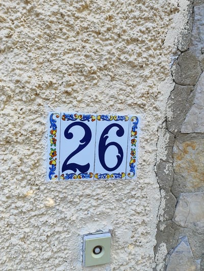 A close-up, eye-level, exterior shot shows a textured, light-colored stucco wall with two decorative ceramic tiles featuring the number "26" in dark blue. The tiles are bordered by a colorful, floral pattern with blue, yellow, and red elements. To the right of the tiles, the wall transitions to rough, grey stone. Below the tiles and slightly to the left, a cream-colored electrical box with a circular button is visible. The scene is part of the facade of a building in Els Poblets, Spain.