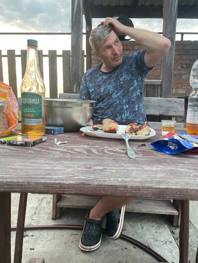 A middle-aged Caucasian man with graying hair and a blue patterned t-shirt is sitting at a wooden table outdoors, holding a black baseball cap on his head with his right hand. He is looking to his right. The table is covered with food and drinks, including a bottle of vodka labeled "КЕДРОВИЦА", a plate of grilled meat, a glass of amber liquid, a pack of cigarettes, and a bag of snacks. The background shows a wooden fence and a brick wall. The man is wearing black Crocs with a red and white trim. The scene appears to be a casual outdoor gathering, possibly in the city of Chulym, Russia, given the context.
