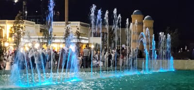 This nighttime scene captures a vibrant public square, likely a tourist attraction in Satkak, Uzbekistan, illuminated by artificial lights. The main subjects are a group of people gathered near a large, illuminated fountain. The fountain features numerous jets of water, some of which are lit with a striking blue light, creating a dynamic and colorful display.
In the background, there are buildings with ornate architectural details, including arched windows and decorative patterns along the facades, suggesting a cultural or historical setting. Two distinctive domed towers, characteristic of Central Asian architecture, are visible on the right side of the frame. The overall atmosphere appears festive and lively, with people strolling, congregating, and enjoying the evening. The time of day is clearly night, with a dark sky above, and the weather seems pleasant, as indicated by the outdoor gathering. The presence of the fountain and the surrounding architecture strongly points to a well-developed tourist area. No visible text is present in the image.