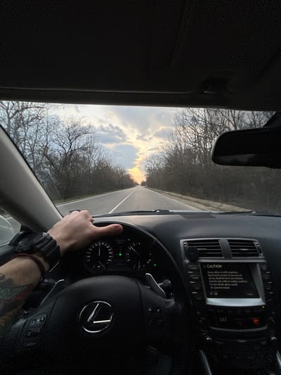 The scene depicts the interior of a Lexus vehicle, observed from the driver's perspective, operating on a straight two-lane asphalt road near Stolnik, Bulgaria. A driver's right hand, featuring tattoos on the forearm, a dark digital wristwatch, and a red string bracelet, grips the steering wheel. The steering wheel prominently displays a Lexus emblem and has paddle shifters visible behind it. The dashboard instrumentation is illuminated, showing the speedometer needle indicating approximately 100-110 km/h. The infotainment screen to the right displays a "CAUTION" message regarding adherence to traffic regulations. Through the windshield, the road extends into the distance, bordered on both sides by dense, leafless deciduous trees. The sky above features scattered clouds, with a bright orange-yellow glow on the horizon, consistent with either sunrise or sunset conditions. A rearview mirror is visible at the top right of the frame.