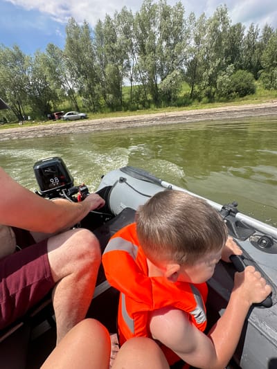 In this upward-angled, close-up shot, a young boy wearing an orange life vest is seated in a boat, his hands gripping a black handle. The boat, a dinghy, is moving through slightly choppy, green water, indicated by splashes around an outboard motor. The motor, with the visible number "8.6" on its casing, is attached to the back of the boat. The boy's hair is brown, and his skin appears wet, suggesting recent water activity.

In the background, to the left of the frame, a shoreline is visible with a line of trees and a patch of grass. Further in the distance, two vehicles, a silver car and a red object (possibly another vehicle or part of one), are parked near the shore. A body of water stretches out towards the horizon, and on the far left, a sliver of blue sky with scattered white clouds is visible. The scene is likely set in the daytime, under fair weather conditions. The overall atmosphere appears to be one of recreation and enjoyment.