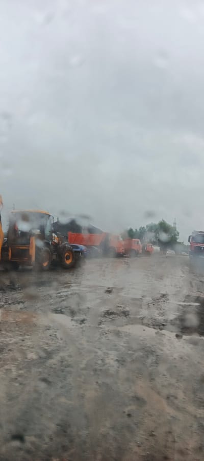 The image depicts a construction site or industrial area in Kabanikha, Russia, under overcast and rainy conditions. The ground is muddy and wet, with visible puddles, suggesting recent or ongoing precipitation.

The main subjects are heavy machinery and vehicles. A yellow and orange backhoe loader is prominently featured on the left side of the frame. To its right, several orange dump trucks are parked. In the background, more trucks and what appears to be a pile of dark material, possibly coal or dirt, are visible. Further in the distance, trees and a power line tower can be seen, indicating an outdoor, likely rural or semi-industrial setting. A white car is also visible in the middle ground, and a red truck is on the far right.

The weather is clearly rainy, with raindrops visible on what appears to be a window or lens through which the photo was taken. The sky is a uniform grey, contributing to the somber mood. The time of day is difficult to determine precisely due to the overcast conditions, but it appears to be daytime.

There is no visible text in the image. The scene conveys a sense of work being carried out despite challenging weather conditions, common in construction or resource extraction environments. The overall impression is one of a functional, industrial setting enduring the elements.