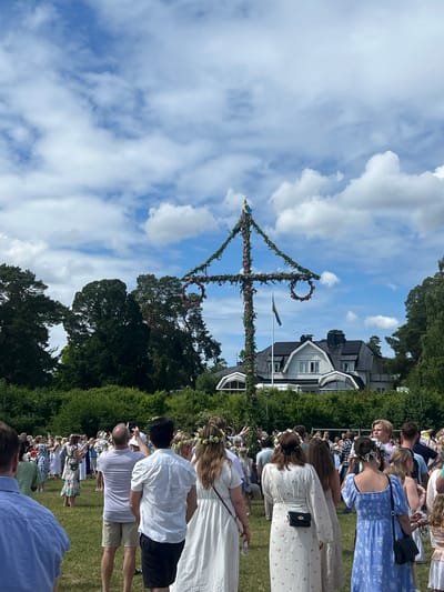 This media file depicts a midsummer celebration in Österskär, Sweden. The scene is outdoors, with a blue sky dotted with fluffy white clouds, suggesting fair weather, likely midday. A prominent maypole, adorned with greenery and flowers, stands tall in the center of the frame. Two Swedish flags are attached to the maypole.

A gathering of people, dressed in festive attire, are seen around the maypole. Many women are wearing white dresses and flower crowns, a traditional element of midsummer celebrations. The people appear to be engaged in or observing an activity associated with the maypole, possibly dancing or singing. The background shows houses with dark roofs and trees, typical of a Swedish residential area. The overall atmosphere seems joyful and communal, characteristic of a summer festival.