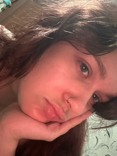 This is a close-up, indoor, waist-up shot of a young person with dark brown hair. They are resting their head on their hand, with their face tilted slightly towards the camera. Their eyes are open and looking towards the viewer, and they have a ring piercing in their nose. Their lips are slightly parted, creating a pouty expression. The background is out of focus but appears to be a wall with a patterned wallpaper. The lighting suggests it is daytime, possibly late afternoon, due to the warm tones and shadows. The setting is likely a home environment. The overall emotion conveyed is one of contemplation or slight melancholy. No text is visible in the image.