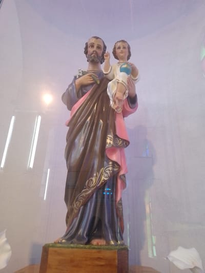 A statue of Saint Joseph holding the infant Jesus stands on a wooden base inside a building, likely a church, in Villa Hayes, Paraguay. Joseph is depicted with a beard and hair, wearing a brown and pink robe with gold-colored detailing. The child Jesus, positioned on Joseph's arm, wears white clothing and holds a blue globe. The statue is located within a glass enclosure. Reflections from light fixtures and windows are visible on the glass, and part of a white flower is visible on the bottom left and right.