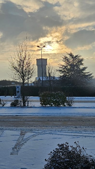 The image depicts a winter scene in Hettange-Grande, France, under an overcast sky with the low sun visible through clouds. The foreground consists of a snow-covered ground with distinct tire tracks in the lower left quadrant. A small, dark, snow-dusted shrub is visible in the lower right. A road surface, appearing partially cleared of snow or slushy, runs horizontally across the mid-ground.
Immediately behind the road, on the left, stands a light-colored electric vehicle charging station. To its right, several bare deciduous trees and dark green evergreen shrubs and hedges are present. A tall, dark grey street light with a dual-lamp fixture is centrally located, silhouetted against the bright sun.
In the mid-background, a large, light-colored cylindrical water tower with a slightly darker cap dominates the skyline, situated behind a row of dark green hedges and a smaller building with a pitched roof. To the right of the water tower, a large evergreen conifer stands prominent. The sun is positioned low in the sky, partially obscured by grey and light orange clouds, casting a warm glow on the upper portion of the scene. No people, specific actions, or direct interactions are visible. The environment indicates cold, snowy conditions with a partially urban or suburban setting.