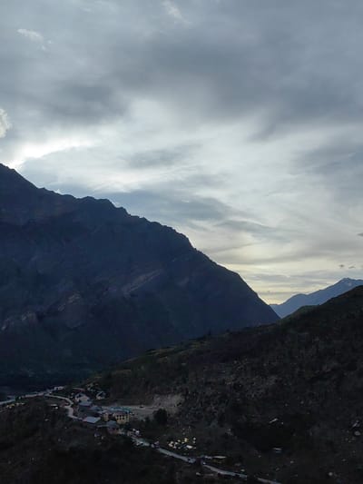 The image depicts a mountainous landscape with a small village nestled in a valley. The sky is overcast with dramatic clouds, hinting at either an approaching storm or recent rainfall. The dominant feature is a large, dark mountain range that occupies the left half of the frame, its rugged slopes and sharp peaks silhouetted against the lighter sky. In the valley below, a scattering of buildings, likely homes and small businesses, form the village of Gushal, India. Roads wind through the village, connecting the structures, and several vehicles are visible. The slopes leading down to the village are covered in sparse vegetation and rocky terrain. The overall impression is one of a remote, high-altitude settlement in a dramatic natural setting.