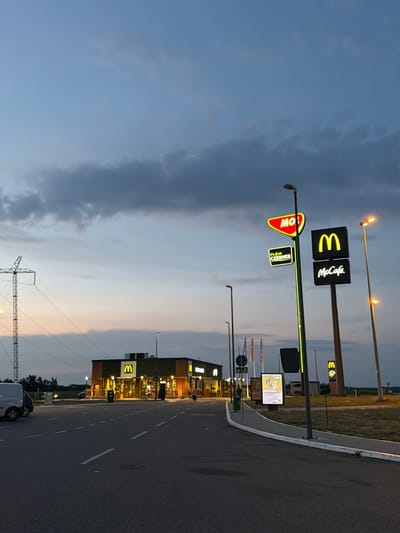 This image captures a McDonald's restaurant located in the city of Ub, Serbia, during twilight. The McDonald's building, illuminated by interior lights, features prominent golden arches on its facade. Outside, several illuminated signs are visible, including a red "MO!" sign, a green "Fresh Corner" sign, and a large black sign displaying the McDonald's logo and "McCafé". A tall utility pole with electrical wires is situated to the left of the McDonald's. The scene includes a paved road with dashed white lines, a sidewalk, and some greenery. The sky is a gradient of blue and gray, with some clouds suggesting the end of the day. A few vehicles, including a white van, are parked near the McDonald's. In the background, distant trees and structures are dimly visible.