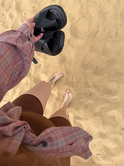 A person's legs and feet are visible on a sandy surface, likely a beach or park. The person is wearing brown shorts and a pink plaid shirt. They are holding a pair of black sandals in their right hand. The sand is light brown and appears somewhat disturbed, with footprints and small debris visible. The lighting suggests it is daytime. The context of Dzerzhinsky, Russia, indicates a potentially urban or suburban park setting with sandy areas.
