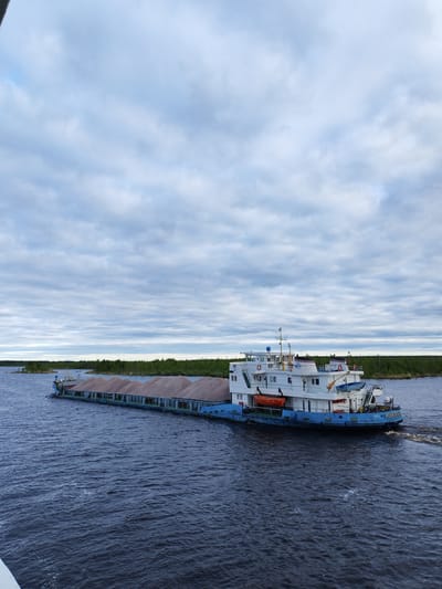 A long cargo barge navigates a wide, dark waterway under a cloudy sky. The barge is laden with a mound of brownish material, presumably gravel or sand, and is marked with “ЯРОСЛАВ МУДРЫЙ” on its lower hull. The barge's main structure is white with a blue lower hull and features multiple windows, life preservers, and a small crane on the top deck. The water surface is rippled, indicating movement. A distant shoreline with green vegetation is visible in the background. The barge's position and the presence of vegetation along the shoreline suggest a setting near a river or lake. It is unknown if this is near the city of Bereg, Russia.