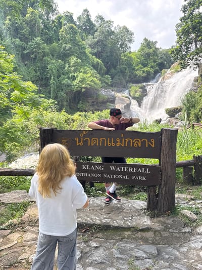 In Ban Namtok Mae Klang, Thailand, a man wearing a maroon t-shirt, black shorts, a black baseball cap with a red floral pattern, and black and red sneakers stands on the right side of a wooden sign. He leans on the sign, gesturing with his right hand towards the waterfall in the background. In front of the sign, a person with blonde hair, wearing a white t-shirt and grey pants with a smiley face pattern, stands facing away from the camera, looking at the waterfall. The sign reads "น้ำตกแม่กลาง" (Namtok Mae Klang) in Thai and "KLANG WATERFALL" and "INTHANON NATIONAL PARK" in English. Behind the sign and the people, a multi-tiered waterfall cascades down rocky terrain surrounded by lush green trees and vegetation. The sky is overcast.