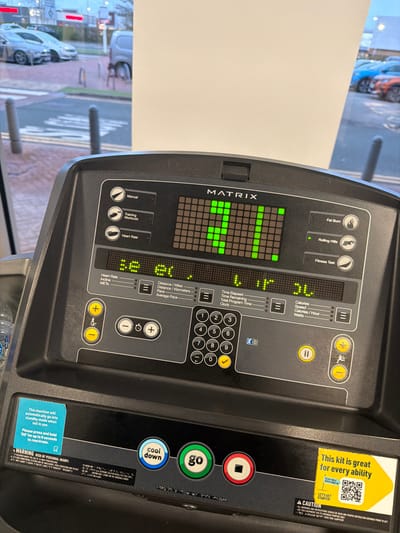 The image depicts the control panel of a black Matrix brand treadmill, positioned indoors facing a window. The central LED display shows the number "30" prominently in green, with additional smaller green numeric readouts indicating "3.0" and "3L" (possibly 3.1 or 3.L). The panel features selection buttons for "Manual," "Training Workouts," "Heart Rate," "Fat Burn," "Rolling Hills," and "Fitness Test." Below these are a numeric keypad (0-9), speed/incline adjustment controls marked with '+' and '-' buttons and icons of a running and walking person, and primary operational buttons: a green "go," a blue "cool down," and a red "stop," alongside a pause button. A blue instructional sticker is affixed, stating, "This machine will automatically go into standby mode when not in use. Please press and hold 'GO' for up to 8 seconds to reactivate." A yellow sticker on the lower right advertises, "This kit is great for every ability" and includes a QR code. A clear plastic water bottle is partially visible in a holder on the left side of the console.
Through the window, an urban street scene in Chester, United Kingdom, is visible. The exterior environment includes a road with distinct pedestrian crossing markings (zebra stripes), several grey bollards, and various parked or moving vehicles, including a silver sedan, a grey van, a blue car, and an orange car. Distant buildings with illuminated signs are present, indicating a commercial area. The overall lighting suggests conditions consistent with dusk or an overcast day.