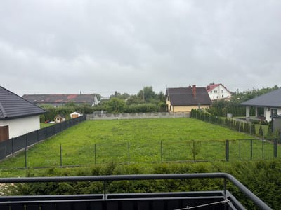 The image depicts a residential backyard in Falenty Nowe, Poland. The scene is viewed from an elevated perspective, likely a balcony or upper floor, overlooking a green, grassy yard enclosed by a dark fence. In the foreground, a dark metal railing partially obscures the view. The yard itself is a large, open expanse of green grass with some patches of weeds.

Beyond the main yard, several houses are visible. To the left, a modern house with a dark, angled roof is prominent. Further in the distance, a long, red-tiled roof house with visible dormer windows stretches across the frame. A yellow house with a dark roof is situated to the right of the red-tiled house. Various trees and dense green foliage line the boundaries of the properties and extend into the distance.

In the background, a concrete fence separates some of the properties. A small, light-colored shed and a small wooden playhouse with a red slide are visible within the fenced yard on the left. The sky is overcast with a thick layer of grey clouds, suggesting a cloudy or rainy day. No people or animals are clearly identifiable in the image. The environment appears to be a suburban or semi-rural residential area.