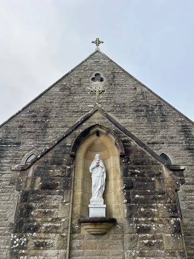 The image shows a low-angle, full-frame shot of the facade of a stone church in Mountcharles, Ireland. A white statue of Jesus, holding a red heart, is positioned in an arched niche in the center of the building. The church's gable is topped with a stone cross. The stone blocks making up the facade are weathered and discolored, with patches of lichen. Small arched windows are visible on either side of the niche. The sky is overcast.