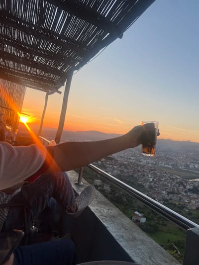 The image depicts a sunset scene overlooking the city of Tétouan, Morocco, from an elevated viewpoint, likely a balcony or terrace. The sky transitions from a clear light blue at the top to vibrant orange and yellow hues at the horizon, where the setting sun creates a strong golden flare above distant mountains.

In the foreground, a person's arm is raised, holding a clear glass containing an amber-colored drink, possibly iced tea or beer, silhouetted against the bright sky. To the right, the legs of another person are visible, clad in dark denim jeans and white sneakers, resting near a grey concrete ledge and a sleek silver metal railing. On the far left, a dark, woven bamboo or reed structure, possibly a canopy or wall, frames the view. Below, the densely packed buildings of Tétouan extend across the landscape under the evening light.

The scene conveys a sense of relaxation and enjoyment during the golden hour. The weather appears clear and pleasant. No visible text is present in the image.