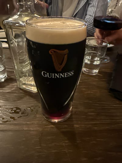 This image, located in Nozawaonsen, Japan, depicts a tall, branded Guinness pint glass positioned centrally on a dark, wood-grained table. The glass contains a dark liquid, topped with a thick, off-white foam head, and exhibits a distinct reddish-purple layer at its base. Condensation or spillage is visible on the glass exterior and forms small puddles on the table surface to its left. In the background, to the left of the Guinness glass, a clear glass decanter or bottle is partially visible. To the right, a small, clear glass tumbler containing water is present. The hand of an individual is shown holding a stemware glass filled with a dark red liquid. A person wearing a light-colored, vertically striped garment is partially visible behind the glasses. A dark, rectangular object, possibly a wallet or menu, rests on the table's far right. No direct interactions are occurring.