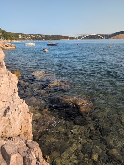 This image depicts a coastal scene in Kraljevica, Croatia, featuring a body of water, a bridge, a shoreline with rocks, and several boats. In the foreground, large, light-brown rocks with visible cracks are prominent. The water in the mid-ground is clear, allowing the rocky seabed to be seen. Several boats are scattered across the water, including a white motorboat and a darker boat. Two individuals are visible in the water; one appears to be swimming, while another is floating on a tube. In the background, a long, arched bridge spans across the water, connecting two landmasses. The shoreline on the left is lined with trees and buildings. The sky is clear and blue, indicating a sunny day.