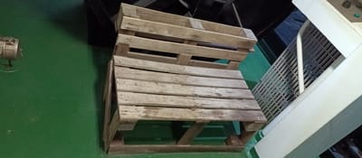 A wooden bench, constructed from two stacked pallets, sits on a green floor. The bench has a seat made of several wooden slats and a backrest formed by the top pallet, also with slats. To the right of the bench, a white metal structure with a slatted surface is visible, partially obscuring a dark background. The overall environment appears to be indoors, possibly a workshop or storage area, located in Al Hamriyah, United Arab Emirates.