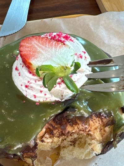 A close-up, overhead view depicts a partially consumed dessert item situated on a light brown paper lining, which rests on a dark brown wooden surface. The dessert features a dark base, extensively coated with a thick, glossy, jade-green glaze. A swirl of white whipped cream is centered atop the green glaze, adorned with a lengthwise-sliced fresh red strawberry half, its green calyx intact. Small, bright red granular sprinkles are scattered across the whipped cream and adjacent glaze. A bite has been taken from the lower right portion of the dessert, exposing a light tan, crumbly interior with darker brown sections. A silver-colored fork with three visible tines is actively inserted into the dessert from the right side. In the upper left corner, the serrated blade of a silver-colored knife is visible on the paper. No individuals are present in the frame. The scene is located in San Phra Net, Thailand.