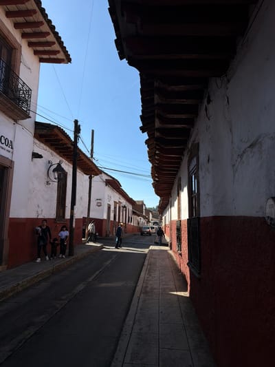 A narrow street stretches between buildings, their walls a mix of white and red. A staircase protrudes from one side. People stand by the wall on the right. Overhead, a bright blue sky is visible between the buildings. Power lines crisscross above. The street is shadowed on one side, revealing details in the textured walls.