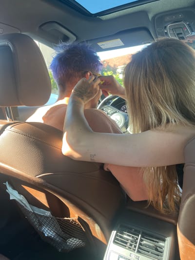 In the city of Pobrđe, Montenegro, a person with vibrant purple hair, viewed from behind, is seated in the passenger seat of a car. Another person, with long blonde hair, is leaning forward from the driver's seat, their right arm extended to adjust something near the purple-haired person's ear. The interior of the car features brown leather seats, a sunroof, and an air vent on the back of the center console. The sunlight streams in through the windows, illuminating the scene and casting shadows. A mesh pocket on the car door contains several white tissues.