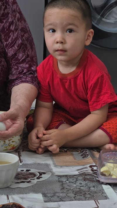 An indoor, waist-up, medium shot captures a young child, likely a boy, with a shaven head and light skin, wearing a red t-shirt and red shorts with a black polka dot pattern. He sits cross-legged on a patterned surface that appears to be a tablecloth. The child looks directly at the camera with a neutral expression. To his left, an adult's arm, belonging to a person wearing a dark purple patterned garment, is visible. The adult's hand holds a white, oval object, possibly an egg, over a bowl with a floral pattern. To the right of the child, a clear plastic container holds sliced yellow food, perhaps butter or cheese. A white mug with a pale yellow liquid, possibly tea, sits in the foreground to the left. The background is dark and out of focus, with a hint of a metallic appliance, possibly a washing machine, on the far right. The lighting is even and direct, with no shadows. The scene suggests a domestic setting, likely a kitchen or dining area, within Mary City, Turkmenistan.
