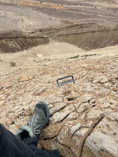 A first-person, downward-angled view captures a steep, arid desert slope in the Arava Tichona Regional Council, Israel. In the lower left foreground, a person's lower left leg, clad in dark pants and a worn grey-blue hiking shoe with light laces, rests firmly on fractured, light brown bedrock. Directly ahead and slightly to the right, a dark, U-shaped metal handhold or step is embedded into the textured rock surface. The immediate and mid-ground are dominated by eroded, porous rock formations, exhibiting a rugged, natural incline. The background reveals an expansive, layered desert landscape characterized by varying shades of tan, brown, and grey geological strata, descending into a valley floor with sparse, scattered vegetation. The scene depicts a challenging mountainous or hilly terrain, likely part of a designated hiking or climbing route.