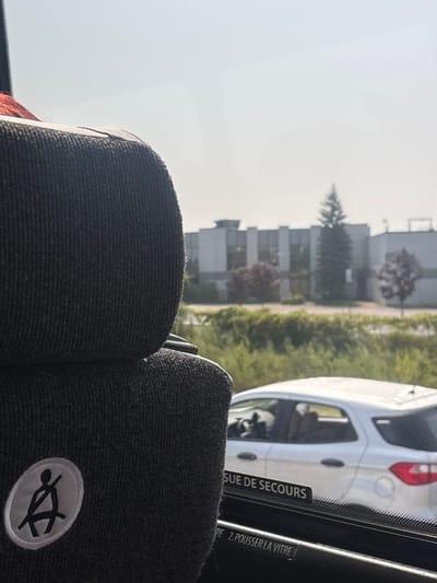 The image depicts an exterior view from inside a vehicle, likely a bus or coach, situated in Boucherville, Canada. In the left foreground, a dark grey, ribbed fabric seat back occupies a significant portion, featuring a circular white emblem with a black seatbelt icon. The black window frame below displays partial white text, including "SUE DE SECOURS" (Emergency Exit) and "RE 2. POUSSER LA VITRE" (Push the Window). Outside the vehicle, a white hatchback car is positioned parallel to the viewer's vehicle. Beyond the car, a strip of dense green foliage acts as a median or shoulder, separating the road from a distant multi-story industrial or commercial building complex. Several trees, including a prominent coniferous tree and smaller deciduous trees, are visible between the foliage and the building. The sky above is bright and appears hazy.