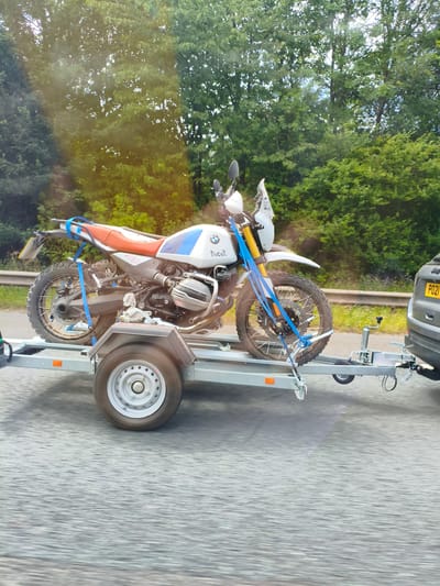 A white BMW R1250GS motorcycle, with blue and white decals and an orange seat, is secured with blue straps to a gray utility trailer. The trailer is hitched to the back of a dark gray Jeep Grand Cherokee SUV, which has a license plate reading "PO21". The vehicles are on a paved road, with a metal guardrail on the left and a dense line of green trees and foliage on the right. The scene appears to be captured from another moving vehicle, as indicated by motion blur in the foreground and background. The image was taken in South Staffordshire, United Kingdom, likely on a rural or suburban road due to the presence of trees and the guardrail. The weather appears to be overcast, with sunlight filtering through the trees, creating some lens flare.