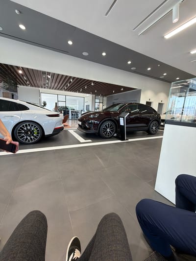 The image captures a modern car dealership interior, likely in Ouahat Sidi Brahim, Morocco, showcasing two high-end SUVs on a polished grey tiled floor. In the foreground, a white Porsche SUV is parked, its rear section and distinctive wheels with bright green brake calipers prominently displayed. To its right, a darker colored Porsche SUV is positioned, its front end visible. A hand holding a dark smartphone is visible on the left side of the frame, suggesting someone is interacting with their device. The dealership features a sleek, contemporary design with a dropped ceiling incorporating recessed lighting and ventilation elements. The background reveals a glass-enclosed area with additional seating and what appears to be a reception desk, further emphasizing the commercial setting. The overall impression is one of a clean, well-lit, and upscale automotive showroom.