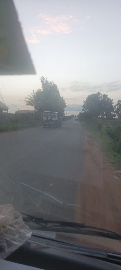 The image shows a road in Mangu Halle, Nigeria. A truck is driving on the road, and another vehicle is further down the road. Trees and vegetation line the sides of the road. The image appears to be taken from inside a vehicle. The sky is overcast.