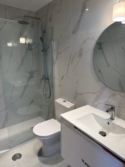 This is a well-lit, eye-level, interior shot of a modern bathroom. The bathroom features marble-patterned tiles on the walls and a simple, light-colored floor. On the left side of the frame is a glass-enclosed shower with a chrome showerhead and handheld shower. To the right of the shower is a white toilet with a chrome flush button. Further to the right is a white vanity with a sink and a chrome faucet. A round mirror hangs on the wall above the vanity, reflecting a light fixture. The image is devoid of people and any discernable action. The setting appears to be a residential bathroom, possibly in a hotel or a newly renovated apartment. Based on the provided context, this bathroom is located in Gondomar, Portugal.