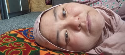 The image is a close-up, tilted, over-the-shoulder shot of a person wearing a light pink hijab with glittery embellishments. The person's face is the primary subject, with their eyes open and looking towards the camera. Their skin appears to have some texture and light makeup. They are lying on a colorful, patterned surface which appears to be a pillow or cushion with intricate red, orange, blue, and green designs. In the background, a blurred grey carpet is visible, and to the left, a dark cabinet or piece of furniture. The overall setting suggests an indoor environment, possibly a home. The image includes a timestamp in the bottom right corner: "21.06.2026 09:08". The location of Chon-Kara, Kyrgyzstan is not visually ascertainable from the image itself.