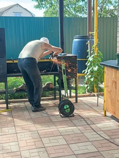 A person wearing a light-colored t-shirt and dark pants is using an angle grinder to cut metal, producing sparks. The scene takes place outdoors on a paved patio, likely in the city of Maslova Pristan', Russia. Behind the person, a green corrugated metal fence is visible, along with a blue plastic barrel and some greenery. There is also a metal structure with shelves where the person is working. A green leveling tool and a black extension cord reel are on the ground near the person. The overall environment suggests a backyard or workshop setting.