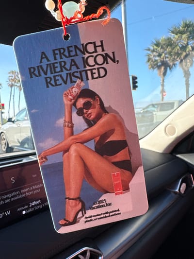 Here is a description of the image:
The image shows a car interior with an air freshener hanging from the rearview mirror. The air freshener has a picture of a woman in a bikini on a blue background with the text "A FRENCH RIVIERA ICON, REVISITED." The air freshener also has the text "Vacation Inc." and "Avoid contact with painted, plastic, or varnished surfaces."
The image also shows a car navigation system with the text "navigation, insert a Mazda" and "Altitude: 24feet" The image background shows a palm tree and cars. Based on the GPS coordinates, the car is located in Huntington Beach, California.
