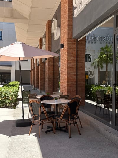 The image depicts an unoccupied outdoor cafe patio situated within the Paseo Milex commercial development in Torreón, Mexico. The foreground features four circular tables with white marble-patterned tops and black bases, each encircled by multiple chairs constructed with dark woven backs and seats, and light-brown frames; a total of at least ten chairs are visible. Small green potted plants are present on two of the tables. To the left, a partially open, light-beige outdoor umbrella with a black base casts a distinct shadow on the light-grey paved ground. Adjacent to the umbrella stands a black A-frame sign displaying white text including "ELLAZ" and "nilba enter ENDLESS LUXURY."

The background is defined by a modern building facade characterized by prominent red brick columns and a dark-grey upper section. Large glass panels forming the building's front reflect elements from across the plaza, including the inverted text "CENTRO DE DIAGNOSTICO HEALTH CENTER," large white circular graphic designs, and two mature palm trees. The scene is illuminated by direct sunlight, resulting in strong shadows. No individuals are visible.