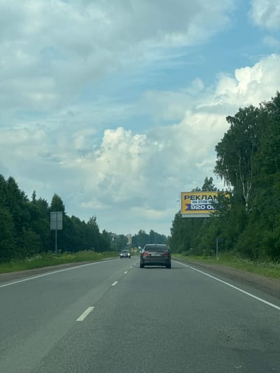This media file depicts a highway scene in Kuzmolovsky, Russia, under a partly cloudy sky. The perspective is from inside a vehicle, looking out towards the right side of the road. The road itself is paved and marked with white lines. Green trees line the sides of the highway, creating a natural border.

Several vehicles are visible on the road. In the foreground, a dark-colored car is driving away from the viewer. Further down the road, other vehicles can be seen, though they are smaller and less distinct.

A notable object in the scene is a large billboard placed on the right side of the road, set against the backdrop of trees. The billboard is yellow and has text on it. The visible text reads "РЕКЛАМ НА ЭТОМ МЕ" and "920 00". Based on the orientation of the billboard and the image, it is likely advertising.

The lighting suggests it is daytime, with the sun visible through the clouds. The presence of lush green trees indicates a warmer season, likely spring or summer. The overall impression is of a typical journey on a highway, with the surrounding natural environment and road infrastructure being the main subjects. There are no people clearly visible, and the focus is on the landscape and the road.