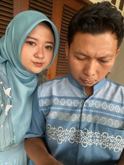 The image displays two individuals, an adult female and an adult male, in a close-up indoor or sheltered setting in Ketegan, Indonesia, against a background of dark brown horizontal wooden slats. The female, positioned on the left, wears a light blue hijab and a matching light blue garment adorned with white butterfly appliques and white pearls on a sheer fabric overlay. She faces the camera with a slight smile, exhibiting visible makeup. The male, on the right, wears a light blue short-sleeved shirt with a mandarin collar, featuring horizontal bands of white geometric patterns and intricate white scrollwork embroidery. His head is tilted downwards, with his eyes closed or averted from the camera. No direct interaction between the subjects is depicted.