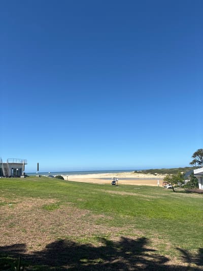 This image depicts a coastal scene under a clear, cloudless blue sky, located in the city of Ngqushwa Nu, South Africa.

In the foreground, a manicured grassy lawn shows variations in color from vibrant green to drier brown patches. Dark shadows from unseen objects are cast across the lower portion of the lawn. No individuals are visible.

To the left midground, a modern two-story building features a light-colored facade, a flat roof, and an external metal-railed staircase leading to an upper balcony. Several dark-colored loungers are positioned on the ground level patio area. Two tall, slender posts and a spherical light fixture stand near the building.

Toward the center-right midground, a small dark vessel is moored on a sandy bank adjacent to a narrow body of water, likely an estuary or river mouth, which extends towards the ocean. A wide expanse of light brown sandy beach is visible beyond the water, backed by vegetated sand dunes.

On the far right, a section of a white building with a wooden deck is partially visible, accompanied by lush green foliage and a small tree.

The background consists of the blue ocean meeting the horizon under the bright, cloudless sky. No people, specific actions, or interactions are observed in the scene.