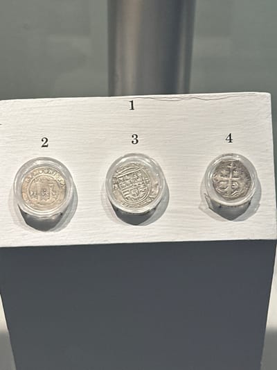 Three historical silver-colored coins are displayed vertically on a textured white surface, each encased in a clear circular plastic holder. The coins are numbered '4', '3', and '2' from top to bottom respectively, with '1' partially visible at the bottom left, suggesting a larger collection.
The top coin (#4) features a prominent cross design, likely a religious or heraldic symbol. The middle coin (#3) displays a detailed, possibly architectural or coat-of-arms design, with faint, illegible text along its rim. The bottom coin (#2) clearly shows a central star or shield motif surrounded by partially visible Latin text, including fragments like 'HISPAN' and 'REX SA'.
The display is set indoors, likely within a museum or historical exhibit in Tepotzotlán, Mexico, consistent with the probable Spanish colonial origin of the coins suggested by the visible text and location context. The lighting creates soft shadows from the coin cases, enhancing their three-dimensional appearance. The scene is static, showcasing the preserved artifacts.