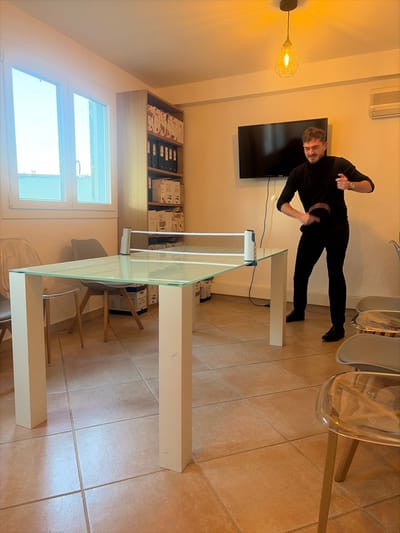 This image depicts an indoor scene in Cagnes-sur-Mer, France. The room features light-colored walls and a floor covered with large, square, light-brown tiles. A large, white-framed window on the left illuminates the room, revealing a light blue sky and distant rooftops outside. A light-colored wooden shelving unit against the rear wall holds numerous black and white binders, along with various white storage boxes. To the right of the shelving unit, a black flat-screen television is mounted on the wall, beneath a white wall-mounted air conditioning unit. A pendant light fixture with a geometric wire frame and an exposed bulb hangs from the ceiling, emitting a warm glow.
Central to the scene is a rectangular table with a clear glass top and sturdy white legs, configured as a table tennis setup with a portable net securely attached across its width. A single male individual, appearing to be in his 20s or 30s, with short dark hair and a beard, is positioned to the right of the table. He is dressed in a black long-sleeved turtleneck shirt, black trousers, and dark shoes. He is actively engaged in playing table tennis, holding a black paddle in his right hand, which is raised and positioned to strike a white ping-pong ball visible in mid-air in front of him. His left hand is slightly raised, and his facial expression indicates focused attention. Several chairs are present: a transparent plastic chair and a grey upholstered chair with wooden legs are on the left side of the table, while two transparent plastic chairs with metal frames are partially visible on the right side, in the foreground.