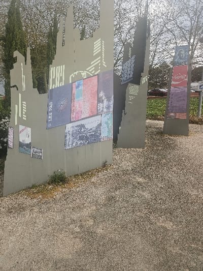 The image displays an outdoor historical or cultural monument located in Avignonet-Lauragais, France. The scene features three primary, vertically oriented panels constructed of a dark grey material, designed with irregular, jagged top edges resembling stylized building silhouettes. These panels are situated on a light-colored gravel ground, with sparse weeds growing at their bases.
Numerous rectangular plaques or posters are affixed to the panels. The leftmost panel showcases several, including one with the text "LES PAS" above a spiral design and another lower down labeled "CATHARE." Other plaques on this panel and the central one display various texts, graphics, and black-and-white photographic or illustrative content. The rightmost, slightly smaller panel features a portrait of an individual at its top, followed by multiple colored plaques, including one with a red background and another with a purple background.
The background consists of a mix of deciduous trees with bare branches, suggesting a late fall or winter season, under an overcast sky. Evergreen trees are visible on the far left. Beyond the gravel, green grass covers the ground. A red vehicle is partially visible behind the rightmost panel, and a white sign with blue text is also partially discernible in the mid-ground. No individuals are present in the depicted scene.