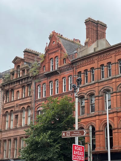 The image shows a street scene with historic red brick buildings on a cloudy day. The buildings have intricate architectural details, including arched windows, decorative brickwork, and chimneys. Vegetation is growing on the rooftops and facades of some of the buildings, suggesting they may be older or less maintained.
In the foreground, there are two signs on a pole. The top sign is a brown directional sign with a pedestrian icon and the text "Tourist Information" pointing to the right. Below it is a red and white sign that reads "ROAD AHEAD CLOSED." A street light or traffic signal with two black cameras is also visible on the pole. A large green tree partially obscures the lower portion of the buildings. The overall impression is of an urban environment with a blend of historical architecture and modern signage.
Based on the signs and architectural style, this scene is likely located in Belfast City District, United Kingdom.
