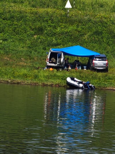 The image depicts a daytime scene near a body of water, likely a river, in Podporozh'e, Russia. The foreground is dominated by the greenish water with ripples, reflecting the environment. Along the bank, there is a grassy area with some vegetation.
On the grassy bank, two silver SUVs are parked next to each other. A bright blue tarp is stretched over them, creating a shaded area. Under the tarp, there appears to be some camping or fishing equipment, including what looks like a small table and chairs. A person is visible sitting under the tarp, facing away from the camera. Several bottles and containers are scattered in the vicinity.
In the water, near the bank, an inflatable boat with a motor is partially submerged. The boat is white with black accents.
In the background, a steep, densely vegetated grassy hillside rises from the bank. There is a white triangular marker with a black pole, possibly a navigational aid or marker, visible near the top of the hill. The overall impression is a scene of outdoor recreation by the water.