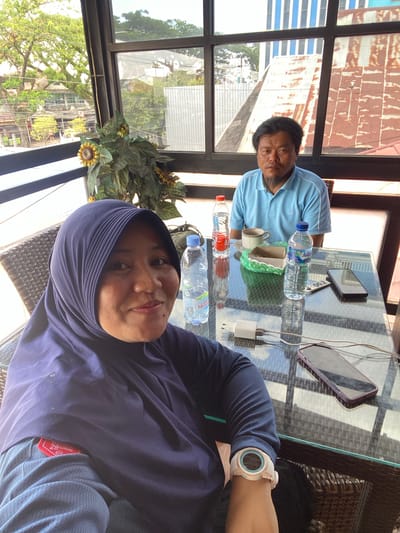 In a sunlit, glass-enclosed patio overlooking a cityscape in Keraton, Indonesia, two individuals are seated at a glass-top table. A woman, wearing a navy blue hijab and a blue shirt with a red tag on the sleeve, is positioned in the foreground, looking towards the camera with a slight smile. A man, wearing a light blue collared shirt, sits opposite her, looking forward with a neutral expression. On the table between them are two bottles of water, a cup of coffee, a small food item wrapped in green paper, a mobile phone, a watch, and a white charging adapter with two cords. The background reveals buildings with corrugated metal roofs and a large sunflower plant in a pot. The time appears to be daytime.