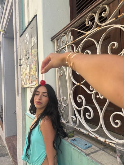 A young woman with long dark hair and closed eyes stands with her head tilted up, a cherry balanced precariously on her forehead. She wears a turquoise tank top with a low neckline. Her eyes are adorned with a small heart-shaped sticker or tattoo under her left eye. Beside her, a metal grate covers a wooden door. A hand with a gold bracelet is visible in the foreground, holding the cherry. On a ledge below the grate, a blue glitter-covered phone case sits next to two plastic cups containing a green liquid and ice. The background shows a textured, pale wall with a dark metal panel covered in stickers, suggesting an urban environment. The architectural style and the presence of graffiti-style stickers on the panel are consistent with the character of Old San Juan.