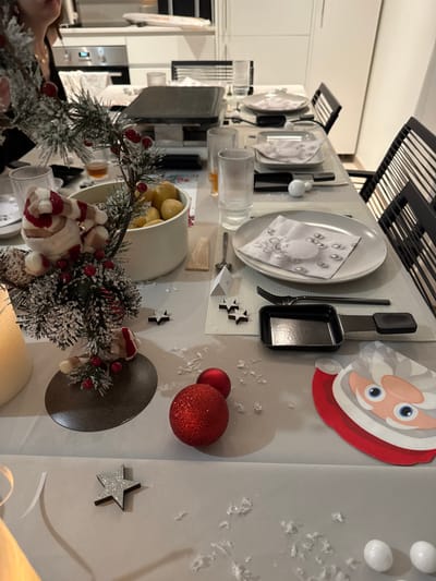 A festive table is immaculately set for a Christmas gathering in a modern indoor setting, likely a dining area or kitchen in Bellevue, Switzerland. The table is adorned with a white tablecloth and individual grey placemats. Each place setting features a light-colored plate with a white napkin, decorated with what appears to be a small white ornament. Alongside the plate, a black fork and a black raclette pan/spatula are neatly placed, suggesting a raclette dinner, a popular Swiss tradition. Ribbed clear glasses are also present at each setting.
The table is extensively decorated with Christmas-themed items. A cheerful Santa Claus cutout is positioned on the right side of the table, near two glittery red baubles. Several small, glittery and plain star decorations are scattered across the table, along with white faux snow or confetti, adding to the winter wonderland ambiance. A prominent pine garland, heavily dusted with faux snow and adorned with red berries and small, festive Santa figures, runs along the length of the table, creating a vibrant centerpiece. A white bowl filled with small, yellow potatoes, typical for raclette, sits among the decorations.
In the background, a modern kitchen with white cabinetry and a black metal shelving unit is visible. A person's arm and hand are partially visible on the far left of the image, suggesting an individual is either seated or assisting with the table setup. The overall scene evokes a warm, celebratory, and inviting atmosphere, ready for a special holiday meal. No readable text is discernible on any of the items.