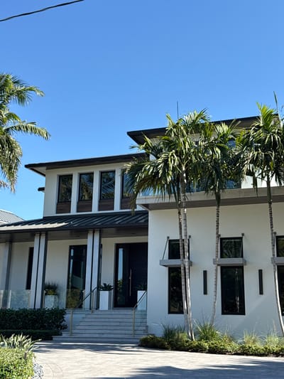 An exterior, ground-level photograph depicts a large, modern two-story residential structure located in Naples, United States of America, under a clear blue sky. The house features white stucco walls, dark grey trim, and a combination of flat and low-pitch roofs, with a darker metal roof section over the main entrance. The upper floor on the left presents four dark-framed rectangular windows, each set above a dark wood-paneled section. The right side of the facade incorporates two vertical pairs of dark-framed windows, with a grey, cantilevered overhang positioned above the lower window in each pair. Vertical dark light fixtures are visible on the white wall sections. The primary entrance consists of wide, light-colored stone steps flanked by clear glass railings with brass-colored handrails. Two prominent dark and light-striped columns frame the tall, dark front door. White cylindrical planters are situated on either side of the entrance at the top of the steps. The property is extensively landscaped with multiple tall palm trees, primarily on the right and left periphery, and various green shrubs and grasses around the foundation. The foreground shows a light-colored paver driveway or walkway. No individuals or specific activities are present. The scene is illuminated by bright daylight, casting defined shadows.